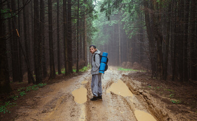 Man in a jacket and gray pants with a backpack on his back stands near a puddle on the road in the mountain forest and looks at the camera.