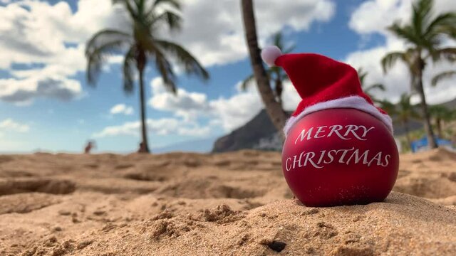 Christmas bomb in Santa's hat on the beach lying on the sand with palm trees and blue sky on the background. Merry christmas from paradise, exotic island. Hawaii, Canary islands, Bali.