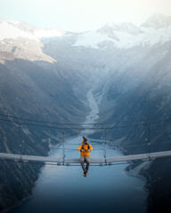 Man in yellow jacket sitting on a suspension bridge over a lake in Olpererhutte, Austria - obrazy, fototapety, plakaty