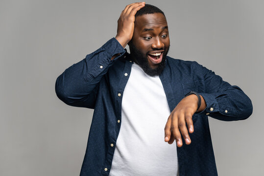 Surprised African-American man wearing casual shirt over t-shirt is late, looks at the hand watch and holding amazed, male office employee forgot about meeting, isolated on grey - Powered by Adobe