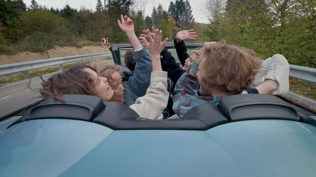 Young People Ride In Car On Road Surrounded By Forest. Beautiful Landscape. They Are Having Fun.They Raise Their Hands And Laugh.
