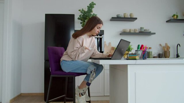Charming positive young woman with prosthetic leg and hand birth anomaly networking on laptop, browsing social media content online while spending leisure in domestic kitchen.