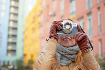 Smiling young woman wearing gloves making photo with retro camera at the city. Focus on the lens.