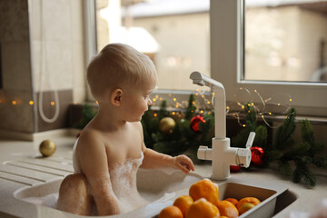 A blonde-haired baby is bathing in the sink in the kitchen against the background of a Christmas tree with garlands and oranges