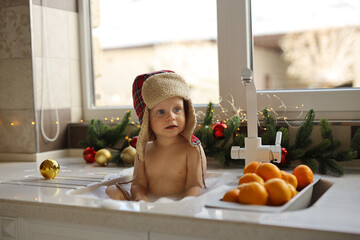 A blonde-haired baby is bathing in the sink in the kitchen against the background of a Christmas...