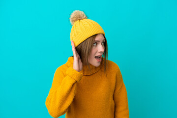 Young English woman wearing winter jacket isolated on blue background listening to something by putting hand on the ear