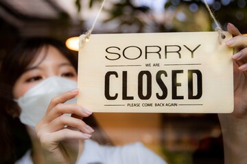 Asian woman wears a mask and turning sorry we are closed sign board at a cafe or restaurant door