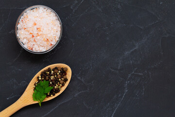 Pink Himalayan salt on the glass bowl and mix of peppers in a wooden spoon on the black background. Top view copy space. Set of various spices for cooking.