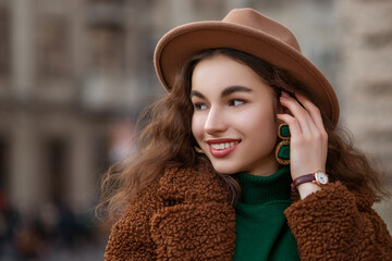 Happy smiling fashionable woman wearing brown fedora hat, wrist watch, big green earrings,...