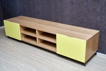 Elegant empty TV stand made of oak wood veneer with bright yellow facade in minimal style near dark cork wall in light living room
