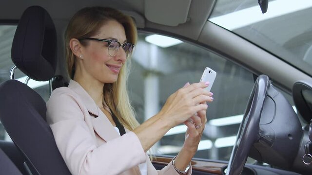 Attractive woman in eyeglasses smiling looking through photos on her smartphone sitting in car, enjoying memories, remembering sweet moments with family, scrolling social networks
