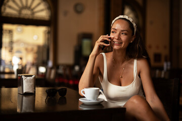Stylish young woman drinking coffee at cafe. Beautiful girl talking to the phone while enjoying in fresh coffee.