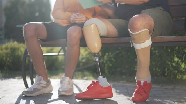 Artificial prosthetic limbs and legs of unrecognizable father and son sitting on bench surfing Internet on tablet in sunshine. Caucasian male amputee and boy resting in park on weekend