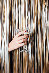 Woman hand is holding martini glass in the middle of Golden foil tinsel strips. Festive background for christmas, new year, holidays or birthday celebration card.