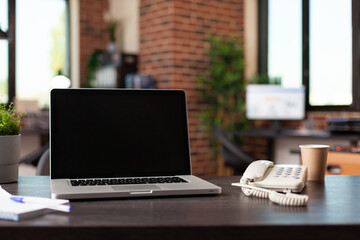 Desk with laptop and landline phone in empty business space. Close up of modern computer, telephone, cup of coffee and office supplies in startup workplace. Workspace decor on table