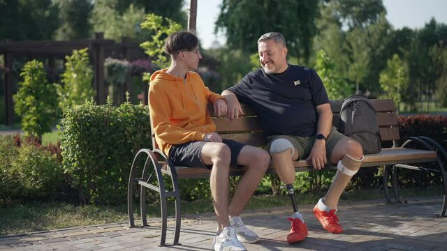 Happy handsome mature amputee greeting teenage boy sitting on bench in sunny park. Wide shot portrait confident Caucasian father with prosthesis and cheerful son meeting outdoors on summer day