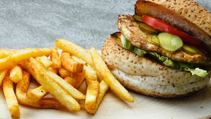 Close-up of tasty chicken burger with french fries, lettuce, tomato and cucumber pickle on a natural wooden tray	