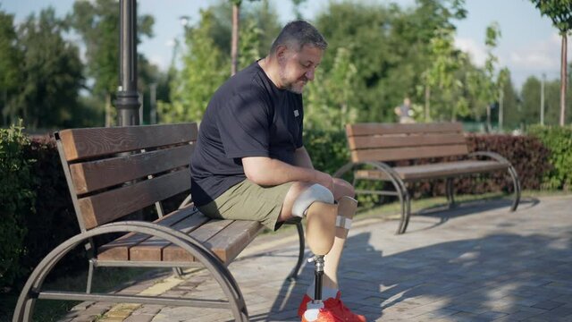 Side view grey-haired male amputee adjusting artificial leg sitting on bench in park on sunny day smiling. Wide shot positive motivated Caucasian mature man enjoying leisure outdoors