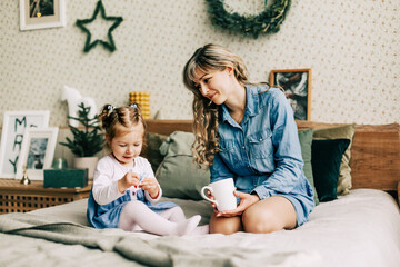 A smiling mother hides a gift for her daughter in a cup, congratulates her on Christmas, sitting on the bed on a winter holiday in a cozy house