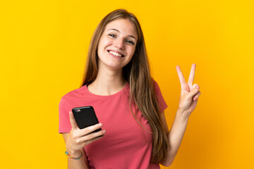 Fototapeta premium Young woman using mobile phone isolated on yellow background smiling and showing victory sign