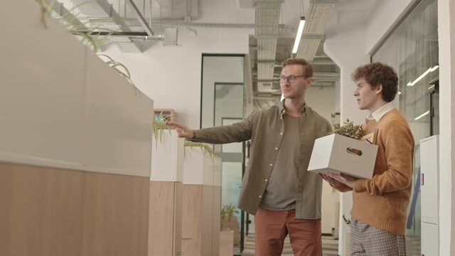 Medium slowmo shot of friendly employer greeting new colleague holding box of his stuff with smile, shaking hands and pointing at his workplace. Young man having first day in office
