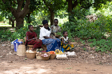 West African farmer's wife and children sitting on the edge of a dirt road waiting for customers to...
