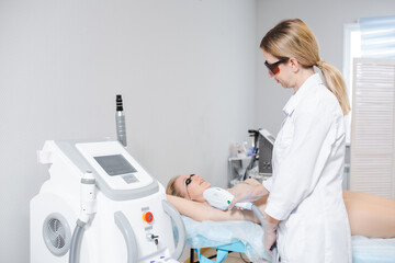 Photo of a modern cosmetology device and a female dermatologist in protective glasses performing a procedure of laser epilation of the armpits of a young girl