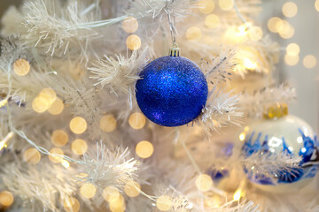 Obraz na płótnie Canvas Blue shiny ball on a white artificial Christmas tree, garland and bokeh. The concept of smart consumption and sustainability. Christmas and New Year 