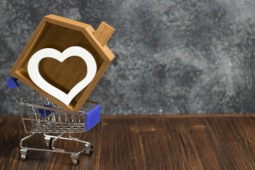 Shopping cart with a wooden house and white heart. Concept for real estate agencies, selling or buying house, loan services and real estate loans. Love house, copy space.