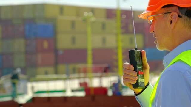 Marine port cargo industry. Senior dock worker in orange hardhat gives instructions to employees against large containers on sunny day close view