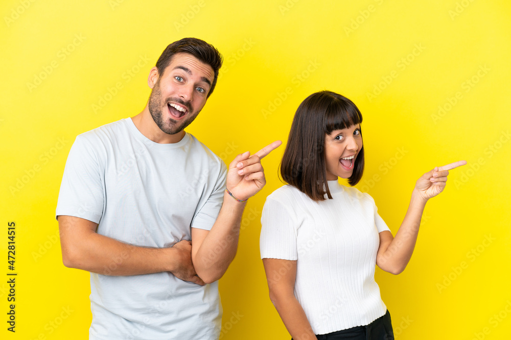 Wall mural young couple isolated on yellow background pointing finger to the side and presenting a product