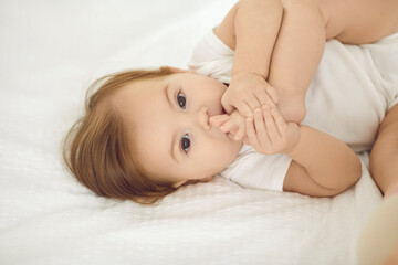 Beautiful carefree little girl of six months lies on white bed linen and plays with her legs. Beautiful baby in white bodysuit pushes hes legs to her mouth looking at camera. Concept of babies.