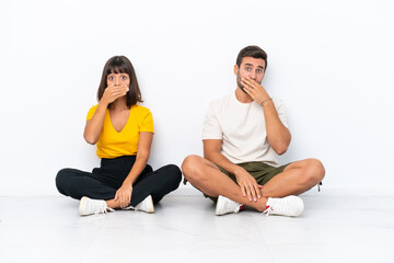 Fototapeta na wymiar Young couple sitting on the floor isolated on white background covering mouth with hands for saying something inappropriate