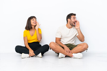 Young couple sitting on the floor isolated on white background shouting with mouth wide open to the lateral
