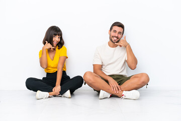 Fototapeta na wymiar Young couple sitting on the floor isolated on white background making phone gesture. Call me back sign