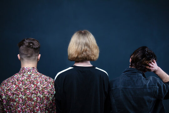 Three Young Men Standing Back To The Camera On Dark Blue Background With Empty Space For Text Or Any Design. Conceptual Picture, Casual Wearing, Blonde And Black Hair.