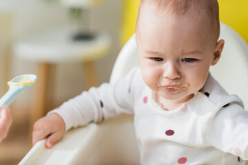 woman holding spoon with food while feeding little son sitting in baby chair.