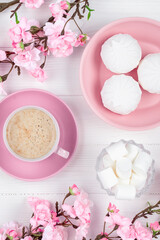 Coffee cup and marshmallow in the morning for breakfast. Spring flowers and dessert on white wooden background. Flat lay. Pastel pink colored card for International Women's Day. Food still life.