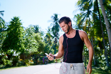 Sport and technology. Young strong man listening music with earphones and smartphone outdoors.