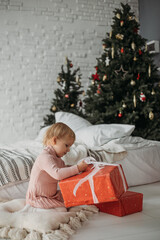 Happy child sitting near the bed and unpacking boxes with her Christmas presents. Happy New Year and Merry Christmas.