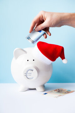 White piggy bank with Santa Claus hat and mans hand puting 20 euro banknote on a blue background. Saving money for Christmas, cash rewards, bonus, sale concept. Vertical image. Selective focus.