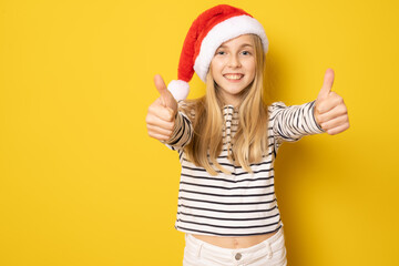 Smiling girl in santa hat showing thumbs up isolated over yellow background.