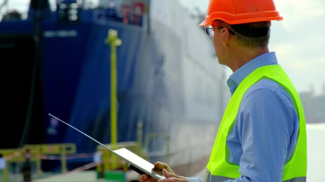 Marine port freight ship loading. Senior customs officer in helmet writes report on tablet standing by open vessel hold in sea dockyard
