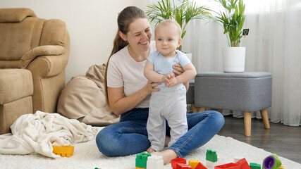 Portrait of happy smiling mother supporting her little baby son standing on acrpet in living room