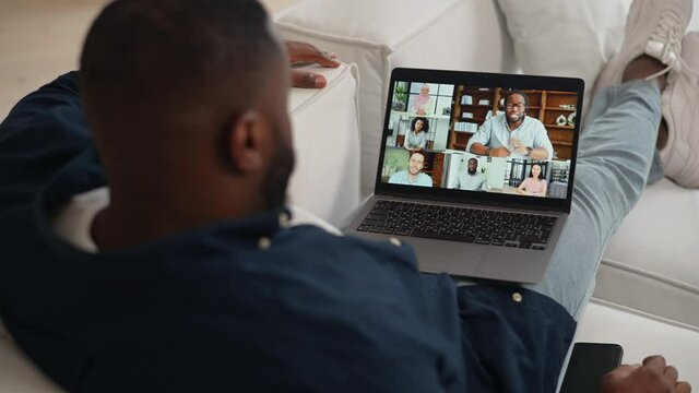 Morning meeting with diverse colleagues. African-American guy using computer app for virtual communication with a group of international people, making video call, greeting, staying at home, back view