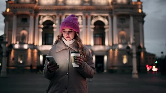 Portrait of attractive blond girl in pink hat texting on her phone. Beautiful young lady at the cold city street. High quality 4k footage