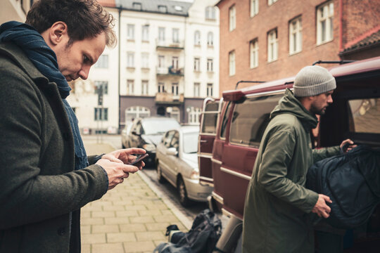 Mature man using smart phone while male friend loading luggage in motor home