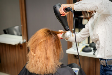 Drying hair in the hair studio. Female hairdresser stylist dries hair with a hairdryer and round brush red hair of a woman in a beauty salon - Powered by Adobe