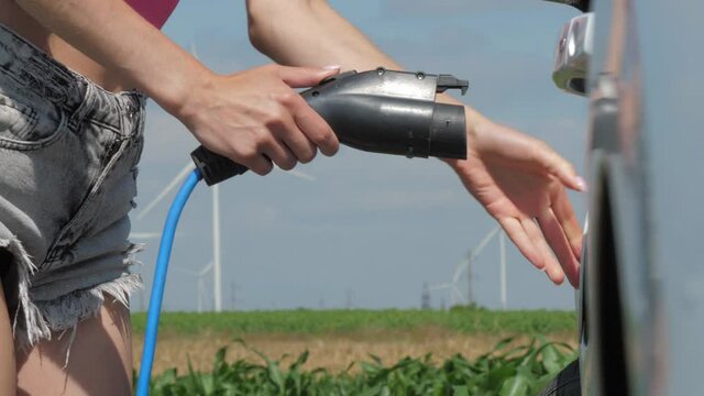 Wind turbines generate alternative energy in corn field. Young woman in shorts charges electric car at windmills station on summer closeup