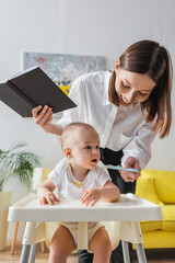 cheerful woman with notebook feeding little son with puree at home.